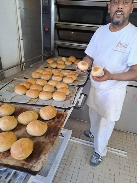 Le boulanger devant le four, un pain à burger dans chaque main, des plaques de pains dorés devant lui