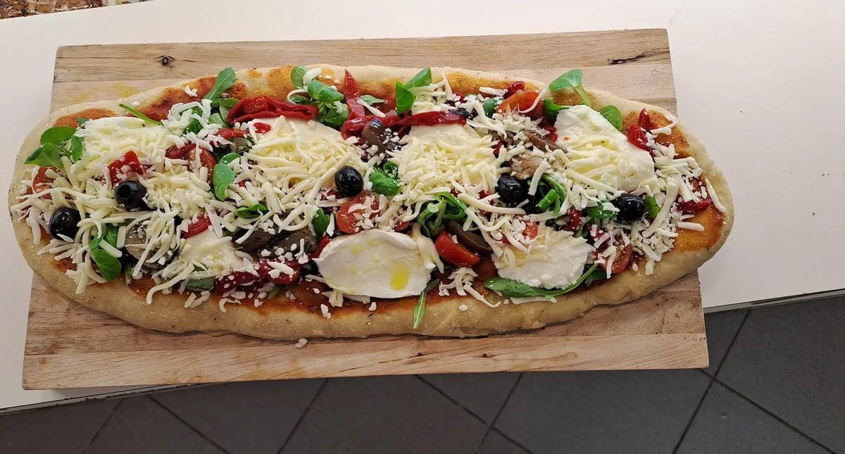 Longue pizza sur une planche de bois : mozzarella, olives noires, roquette, poivrons et fromage râpé
